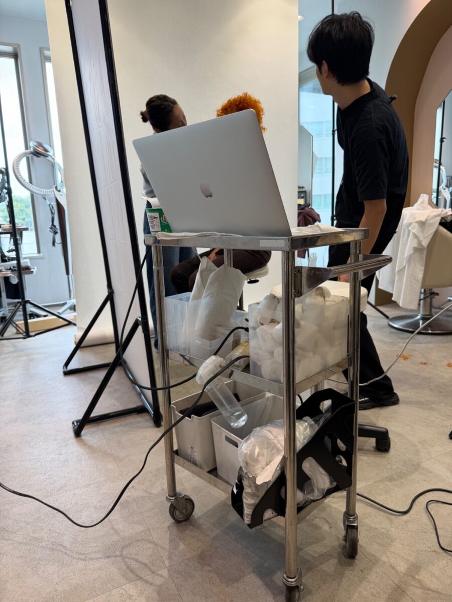 美容室内でオレンジ系カーリーヘアのモデルを撮影するスタッフとMacBookでデータ確認する様子